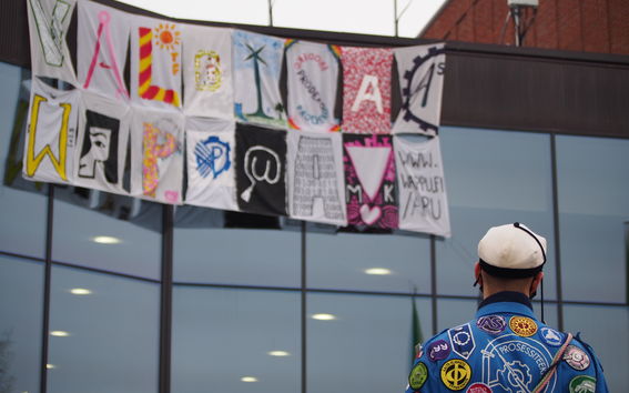 Kuvassa Väreen ulkoseinälle ripustettuna lakanat, joille on maalattu teksti "Valoisaa Wappua". Lakanaa katsoo henkilö opiskelijahaalarissa ja teekkarilakissa.