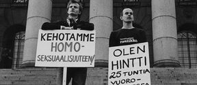 Historiallisessa kuvassa kaksi henkilöä eduskuntatalon portailla pitävät kylttejä, joissa lukee "kehotamme homoseksuaalisuuteen" ja "olen hintti 25 tuntia vuorokaudessa".