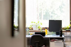 Desk in front of a window with laptop, screen, water bottles, pencils, papers and a plant on it.