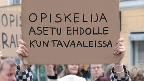 Kuvassa mielenosoituskyltti jonkun käsissä. Pahvisessa kyltissä lukee "opiskelija, asetu ehdolle kuntavaaleissa"