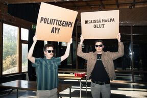 Edustajistovaalit 2025 kampanjakuva jossa kaksi opiskelijaa Smökissä pitää mielenosoituskylttejä, joissa tekstit "poliittisempi AYY" ja "bileissä halpa olut"