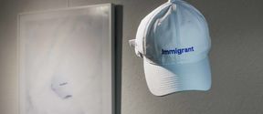 White cap with blue embroidered text with the word "immigrant". In the background a framed poster with a photo of the same kind of cap.