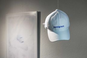 White cap with blue embroidered text with the word "immigrant". In the background a framed poster with a photo of the same kind of cap.