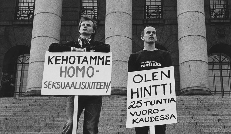 Historiallisessa kuvassa kaksi henkilöä eduskuntatalon portailla pitävät kylttejä, joissa lukee "kehotamme homoseksuaalisuuteen" ja "olen hintti 25 tuntia vuorokaudessa".